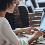 Une femme concentrée sur son travail devant un ordinateur portable. Ses cheveux bouclés sont mi-longs et elle semble être dans un environnement de bureau. Ses mains sont posées sur le clavier, suggérant qu'elle est en pleine rédaction ou navigation.