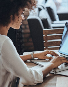 Une femme concentrée sur son travail devant un ordinateur portable. Ses cheveux bouclés sont mi-longs et elle semble être dans un environnement de bureau. Ses mains sont posées sur le clavier, suggérant qu'elle est en pleine rédaction ou navigation.